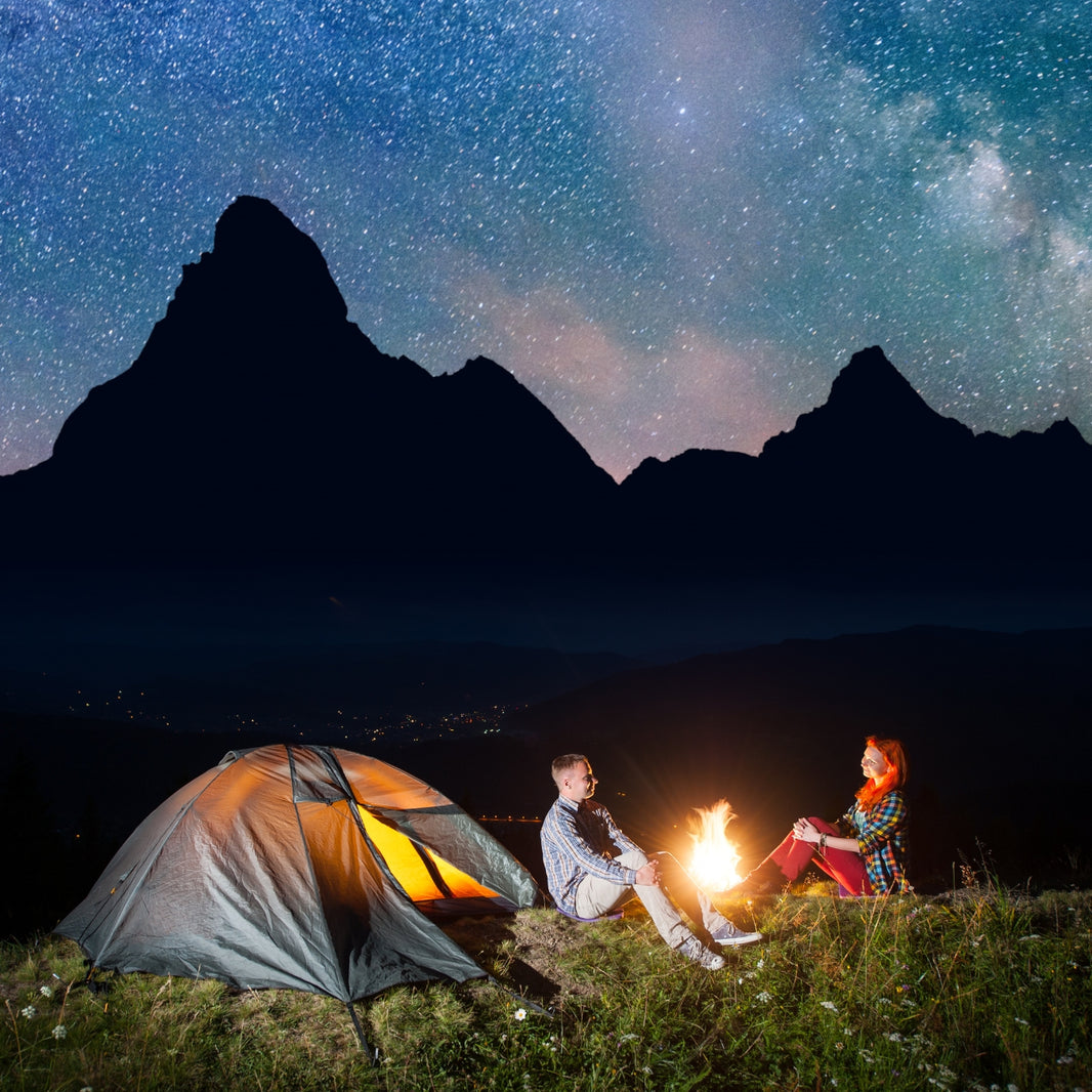 Two people sitting by a campfire under a starry sky with mountains in the background.